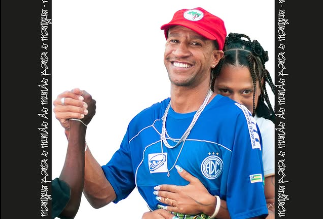 A imagem mostra três pessoas, sendo dois homens e uma mulher, em pé perto uma da outra. A pessoa no centro usa uma camisa esportiva azul com as letras "ADC" e um logotipo, além de um boné vermelho do MST. A pessoa à esquerda aperta a mão do Bidu pessoa ao centro, enquanto a mulher atrás de Biduo abraça por trás, com as mãos descansando em sua cintura.